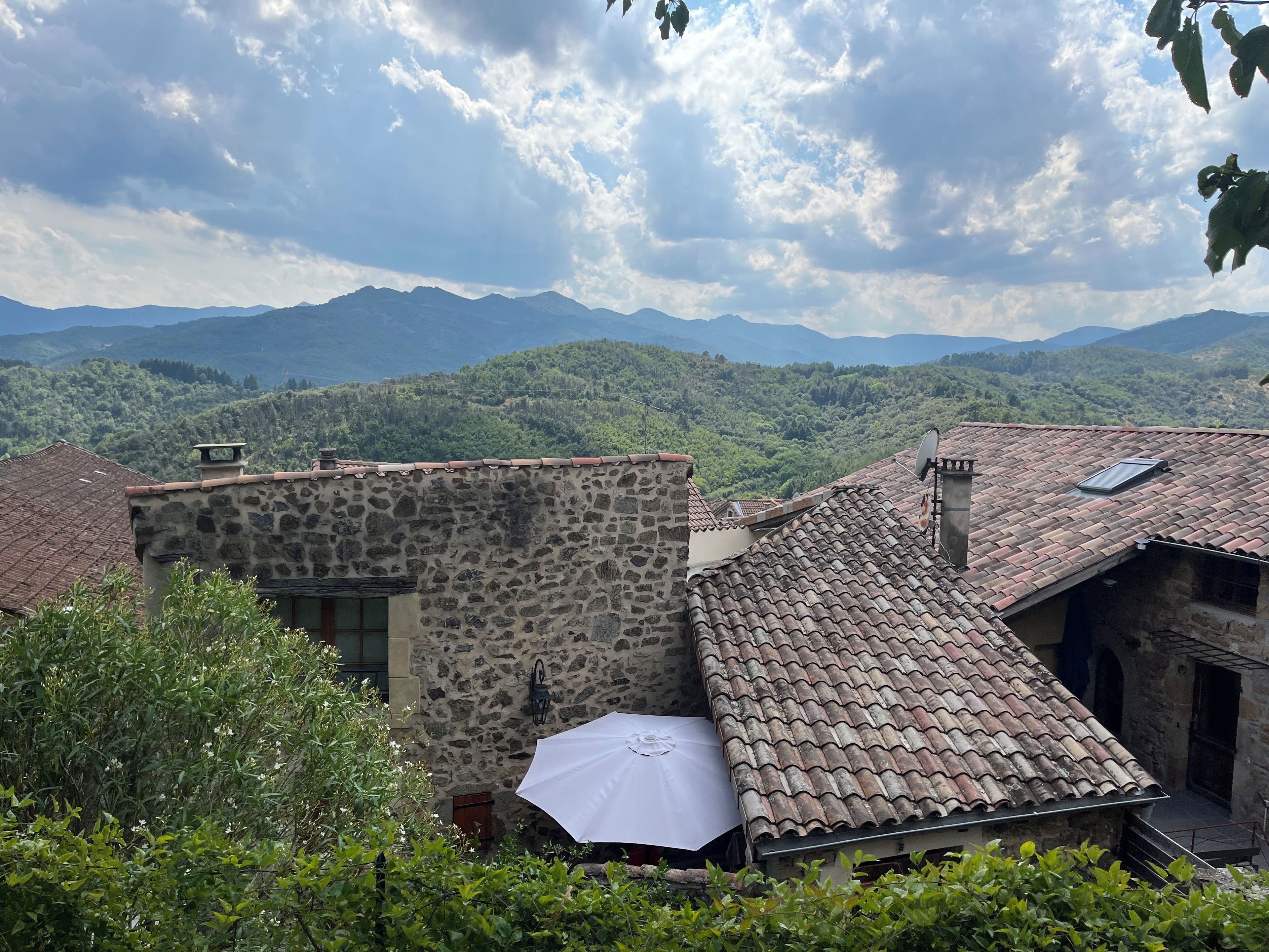 Un edificio de piedra con tejado de baldosas, rodeado de vegetación y montañas al fondo.