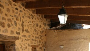 A rustic stone-walled room with a wooden table and benches, a metal shelf, and a hanging lantern.