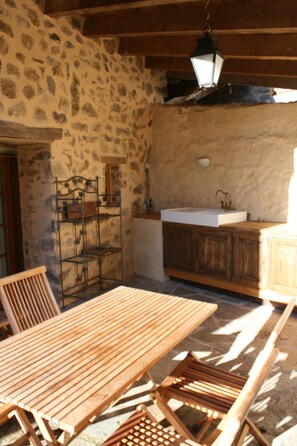Una habitación rústica con paredes de piedra, una mesa de madera y bancos, un estante metálico y un farol colgante.