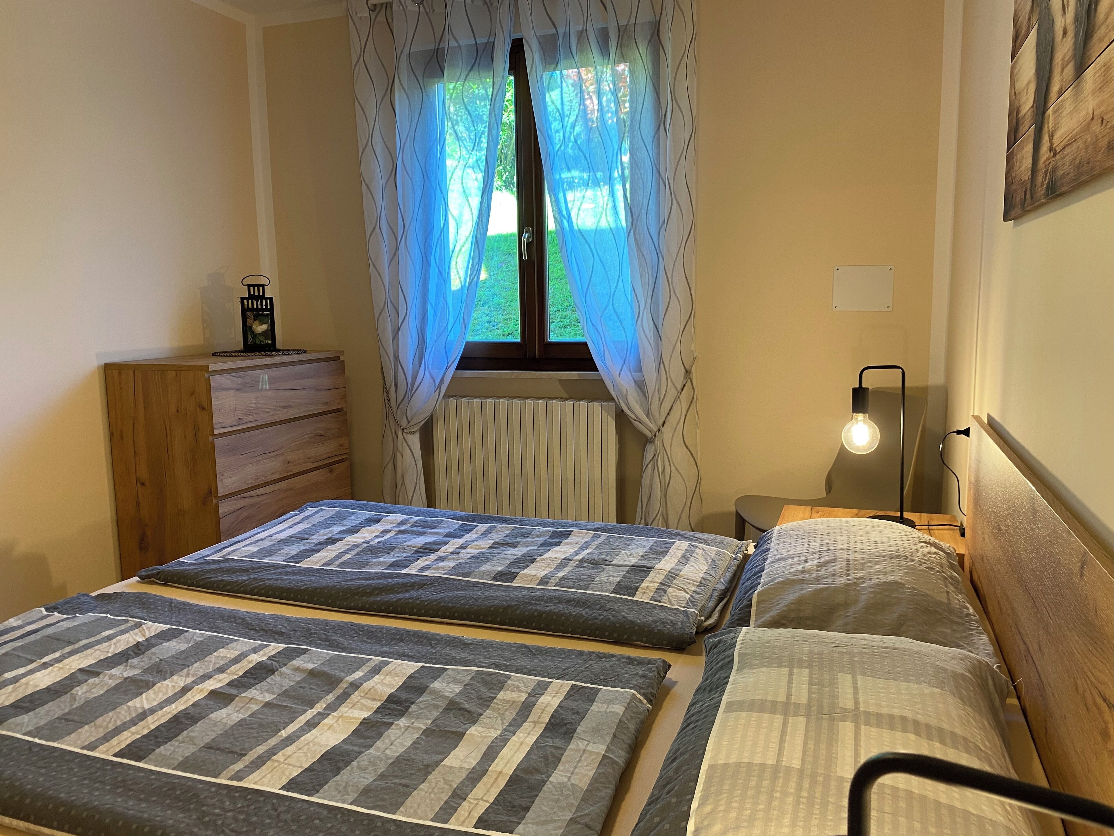 Un dormitorio con dos camas, un aparador de madera, un radiador, una lámpara y una ventana con cortinas traslúcidas.