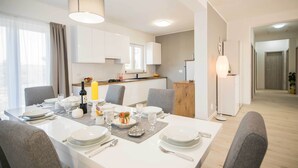 Un espace repas moderne avec une table blanche dressée pour quatre personnes, une cuisine en arrière-plan et un couloir menant à une autre pièce.