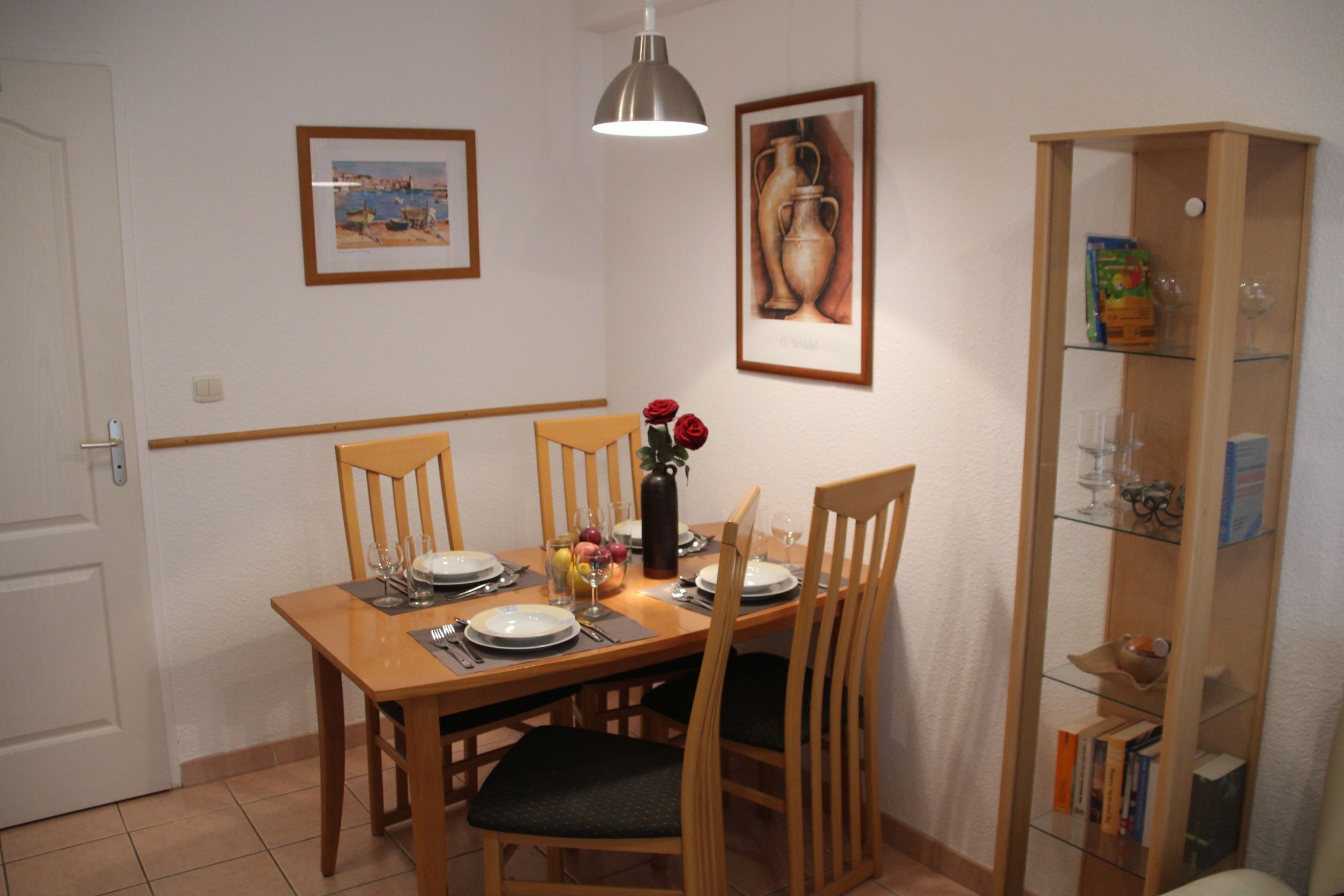 Un comedor con una mesa de madera dispuesta para cuatro, sillas, un jarrón con flores y cuadros en la pared.