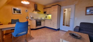 Una cocina compacta con estufa, microondas y armarios, un comedor con mesa y sillas, y una pequeña zona de estar con sofá.