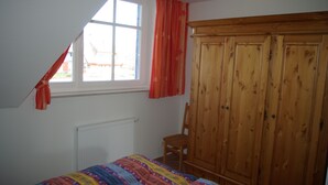 A bedroom with a sloped ceiling, a window with red curtains, a wooden wardrobe, a bed with a colorful blanket, and a radiator. - Darss-Wieck, living room-kitchen-bedroom-balcony, sauna, 2- 3 persons, bicycle shed (Wieck)