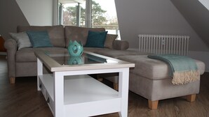 Una sala de estar moderna con una mesa de centro de vidrio, un sofá con cojines verde mar y un pequeño otoman con una manta.