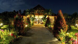 Een hotel met een traditionele architectuur, herkenbaar aan een opvallende ingang, een goed verlicht pad en weelderige beplanting.
