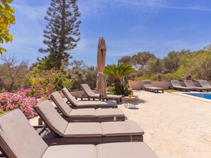 Un espace aménagé au bord de la piscine, avec des chaises longues et une petite table, entouré de plantes en pot et d’arbres.
