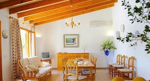 A cozy room with a wooden ceiling, a sofa, a dining table with chairs, a TV, and a potted plant.