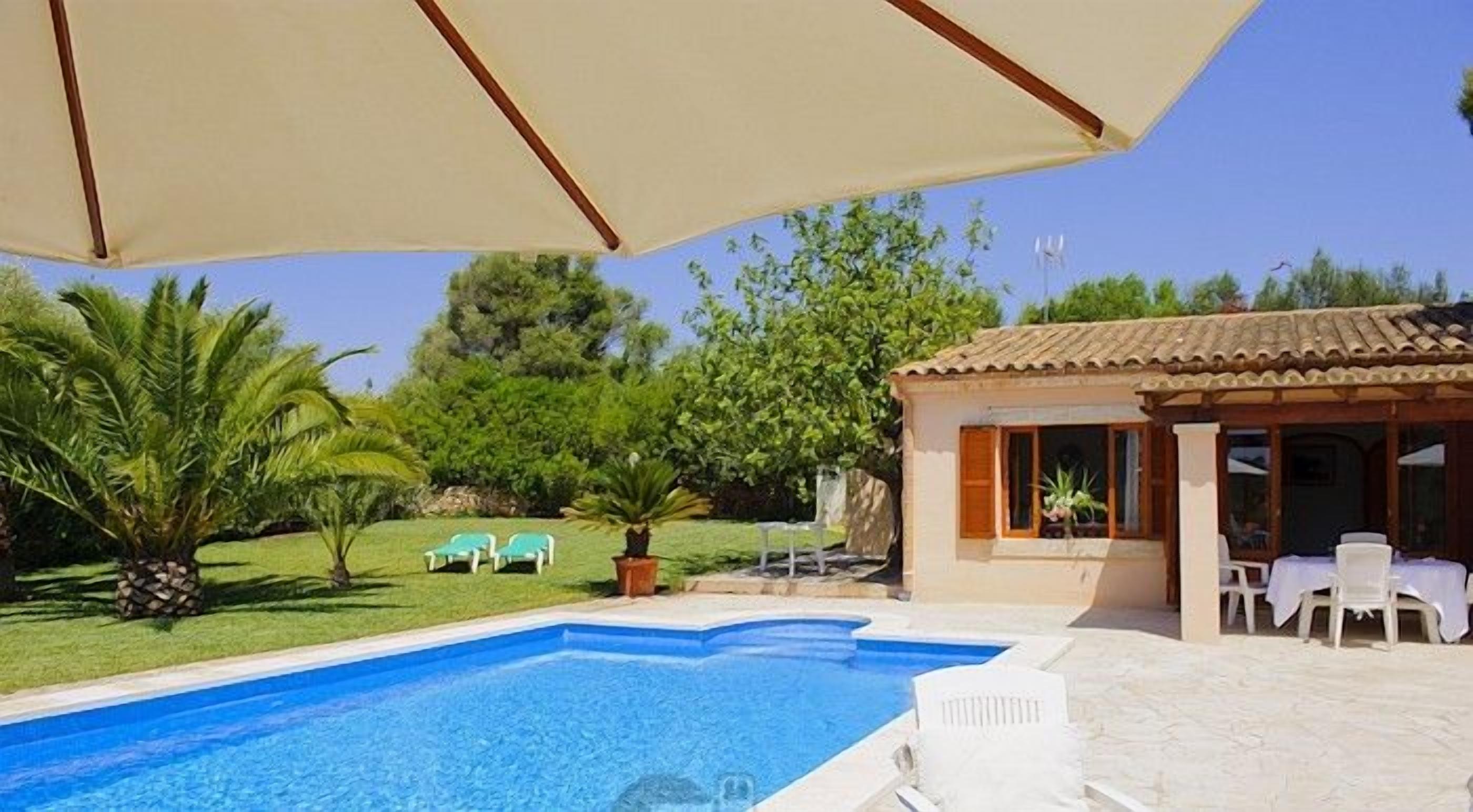 Piscina con sillas de descanso y sombrillas, rodeada de vegetación y un pequeño edificio con mesas al aire libre.
