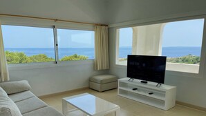 Een woonkamer met een bank, een salontafel, een televisie op een witte standaard en grote ramen met uitzicht op de zee.
