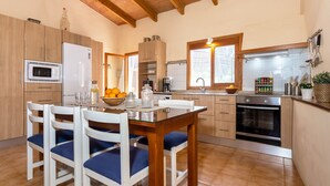 A modern kitchen with wooden cabinets, a built-in oven, and a dining area with a glass table and white chairs. - Finca Son Rebassa (Sant Joan)