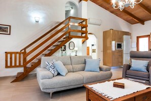Een woonkamer met een houten trap, een grijze bank, een houten salontafel en een keuken met apparatuur.