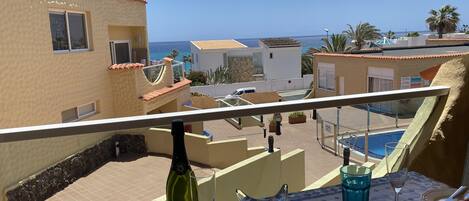 A balcony with a table set for two, a bottle of wine, and a glass. The view includes a pool and buildings with a coastal backdrop.