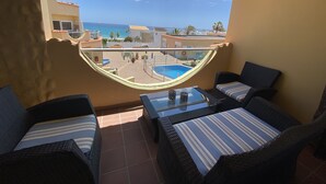 Un balcón con mobiliario de mimbre, una mesa de vidrio y vistas a una piscina y edificios al otro lado del agua.