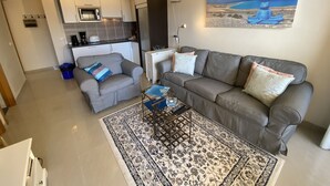 Una sala de estar moderna con un sofá gris, una mesa de centro y una alfombra estampada. Se ve una cocina con armarios blancos y un microondas al fondo.
