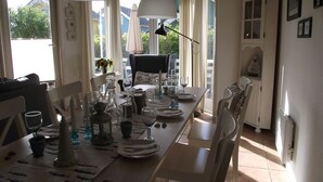Ein Essbereich mit einem langen Tisch, der für eine Mahlzeit gedeckt ist, Stühlen, Hängeleuchten und Blick ins Freie.