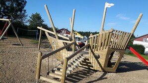 Eine Holzspielanlage mit Rutsche und Kletterelementen, umgeben von einem eingezäunten Bereich mit Sandboden.