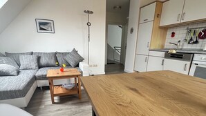Un salon moderne avec un canapé gris, une table basse en bois et un coin cuisine doté de meubles blancs et d’un plan de travail.