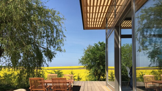 An outdoor deck with wooden furniture, a pergola, and a view of a yellow field.