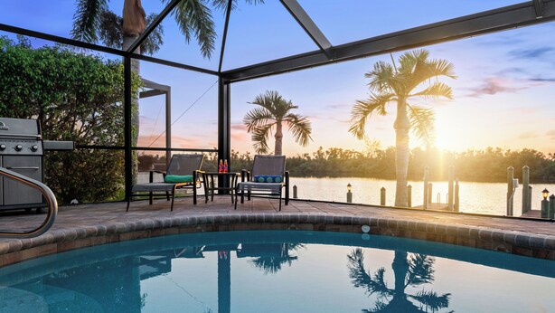 Un espace piscine offrant une vue sur le coucher de soleil sur l’eau, entouré de palmiers et agrémenté de mobilier d’extérieur.