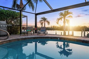 Un espace piscine offrant une vue sur le coucher de soleil sur l’eau, entouré de palmiers et agrémenté de mobilier d’extérieur.