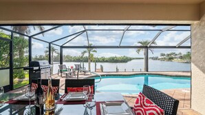 Un coin repas avec une table en verre, des sets de table rouges et une vue sur un lac et des palmiers.