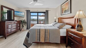 Une chambre à coucher comprenant un lit, une commode, une table de chevet, une lampe, un ventilateur de plafond, une fenêtre avec des rideaux et un tableau accroché au mur.