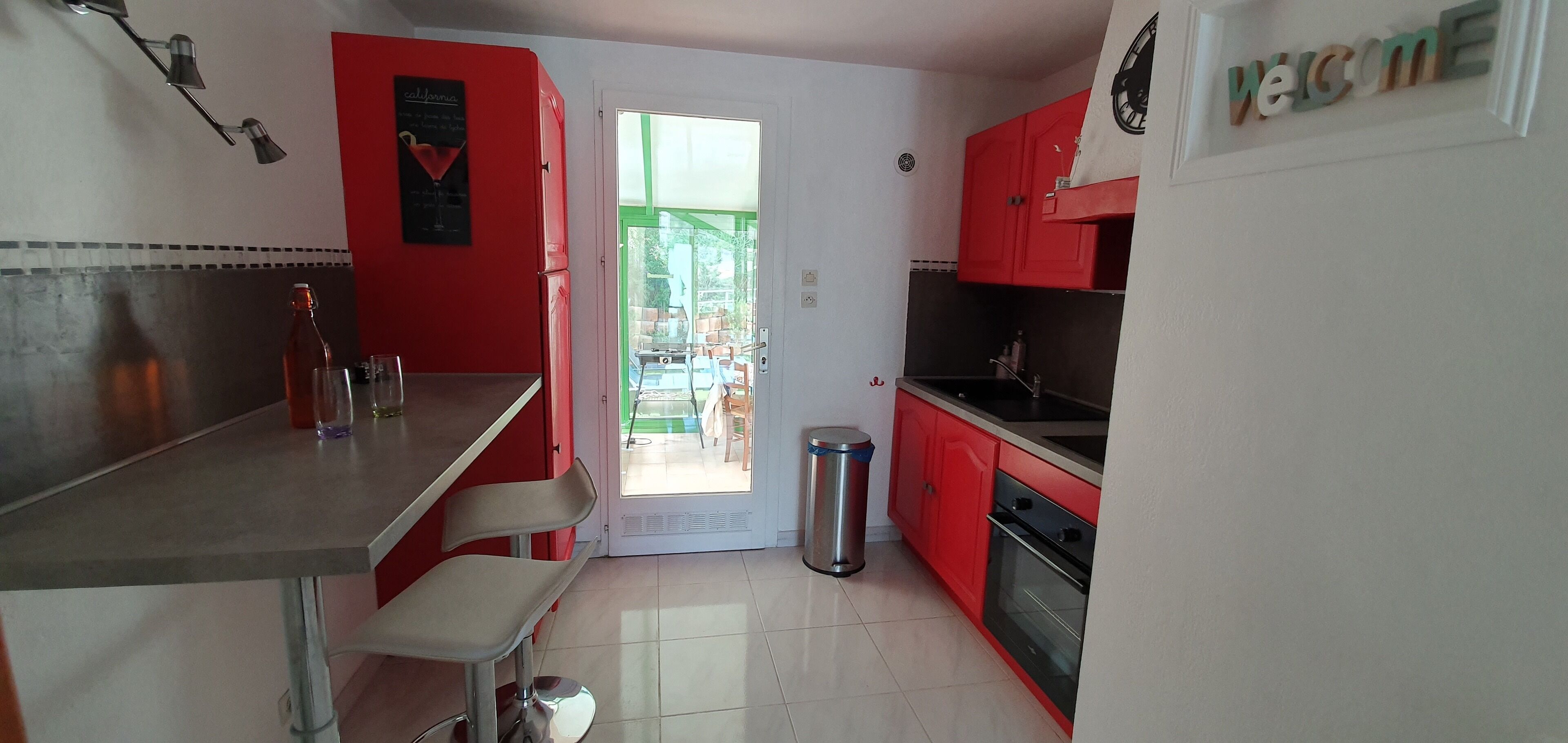 A compact kitchen with red cabinets, a stainless steel countertop, and a small trash bin.