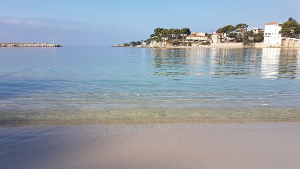 Ein Strand mit klarem Wasser, einer felsigen Küstenlinie und Gebäuden an einem Hügelhang.