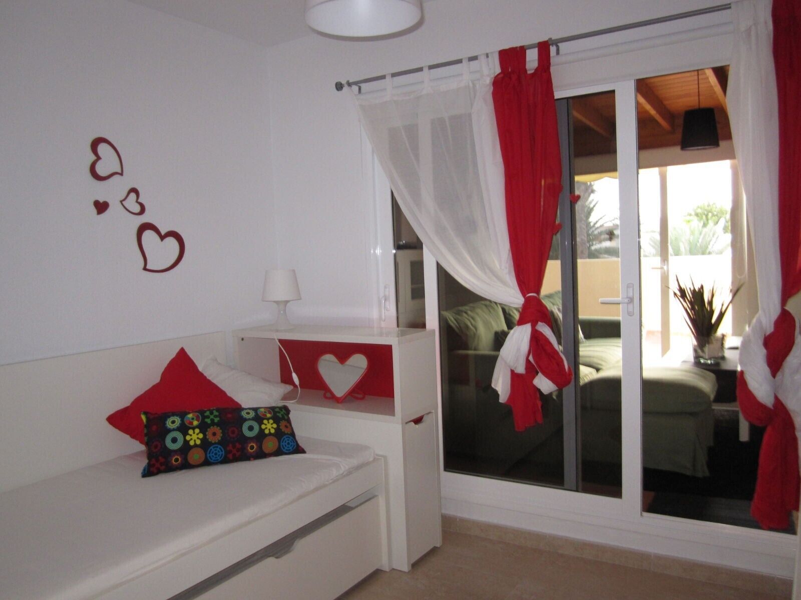 Un dormitorio con cama, mesita de noche, ventana con cortinas y un muro decorado con corazones.