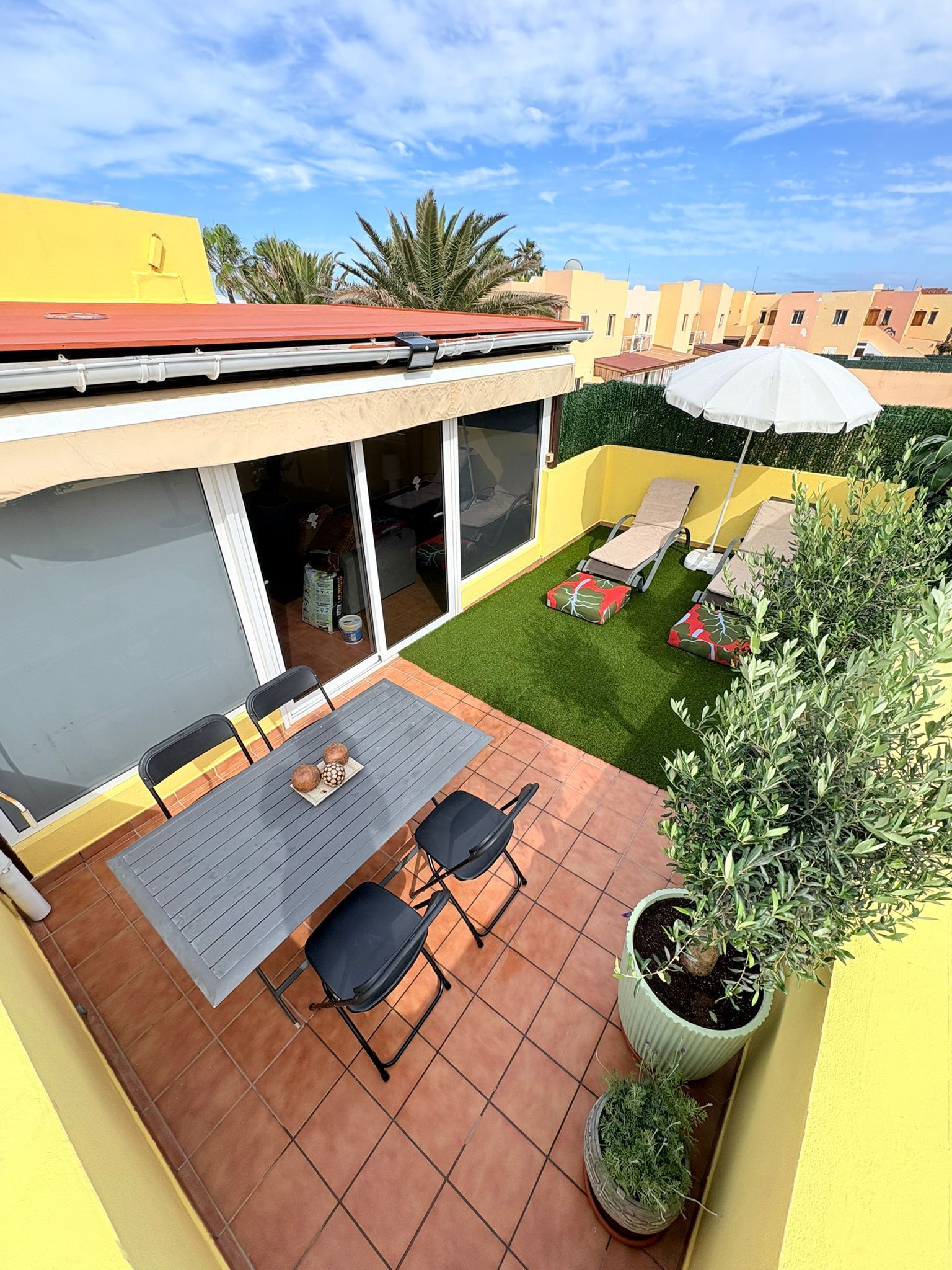 Une terrasse avec une table et des chaises, un espace couvert avec une porte coulissante et une petite pelouse.
