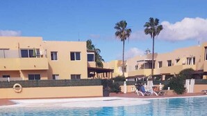 A swimming pool with clear blue water, surrounded by buildings with balconies and palm trees. - Cozy apartment in a well-kept complex with pool, only 40 meters to the beach (Corralejo)