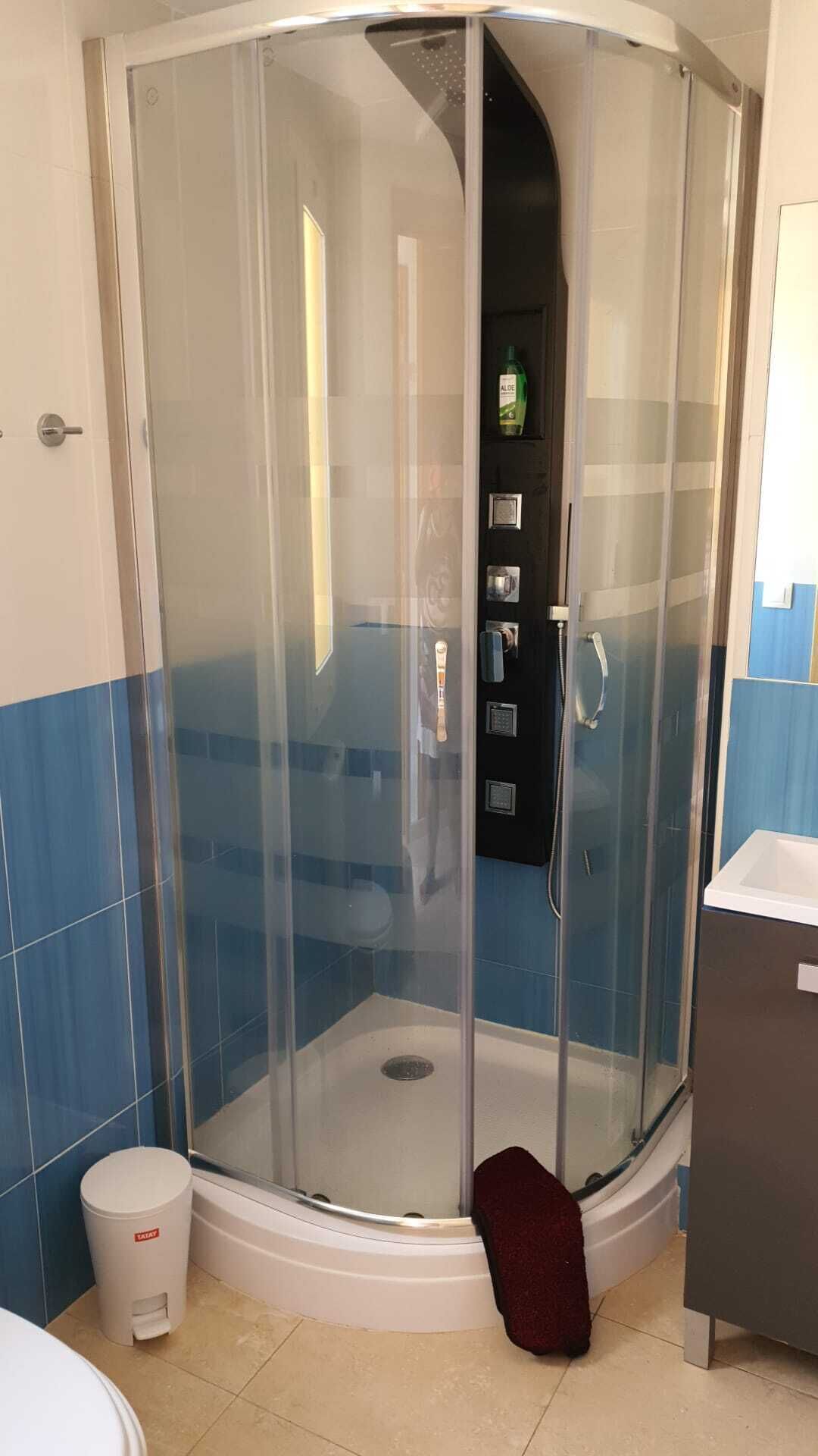 Une douche d’angle avec un pare-douche en verre, une poubelle blanche et un tapis de bain rouge foncé.