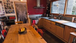 A kitchen with wooden cabinets, a large island with a bowl of fruit, a red wall, and a window with a view of the outside. - WOODEN HOUSE ON THE GULF OF MORBIHAN (Crach)