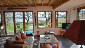 A spacious living room with a large window overlooking a lake, a wooden ceiling, and a cozy seating area with a sofa and armchairs. - WOODEN HOUSE ON THE GULF OF MORBIHAN (Crach)