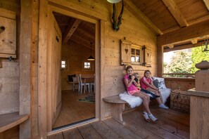 Een houten chalet met een gezellig interieur, met een bank waarop twee personen zitten, een houten kast met een hartvormig raam, en een eethoek die door een open deur zichtbaar is.