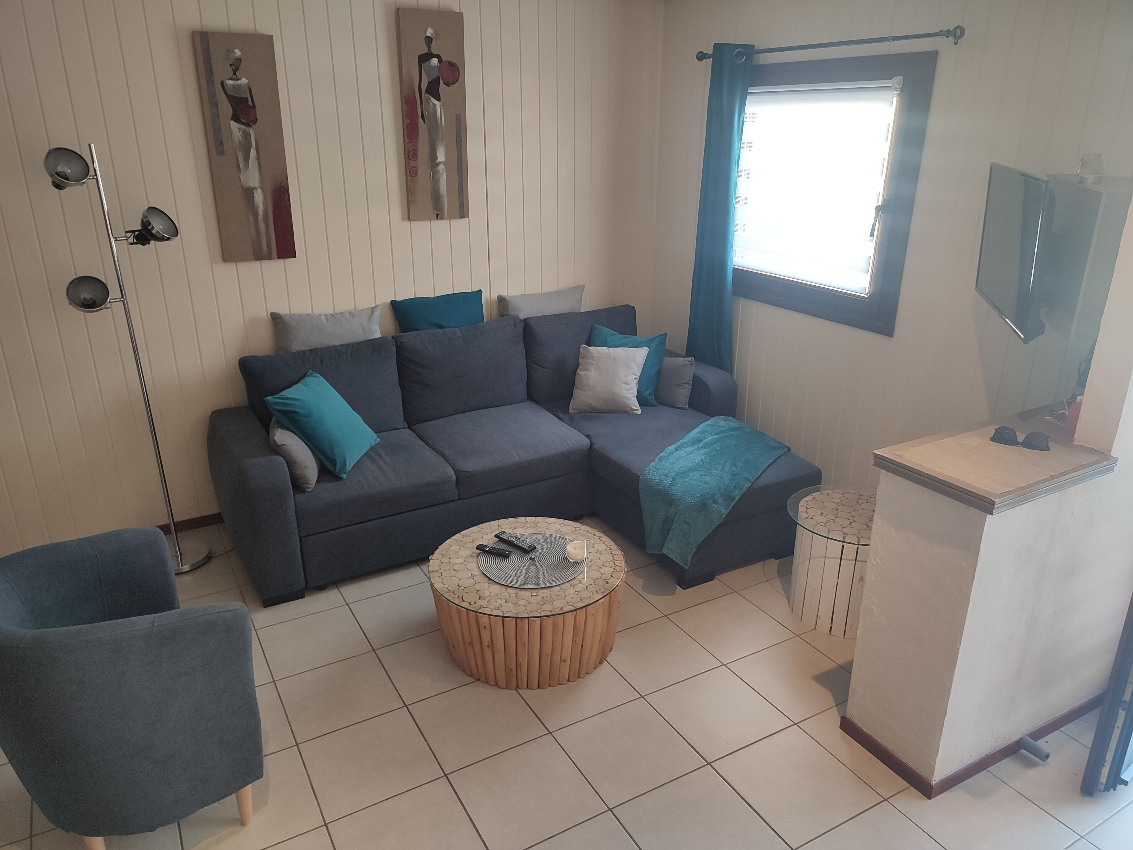 A living room with a grey sectional sofa, a round wooden coffee table, a TV on a stand, and a window with blue curtains.