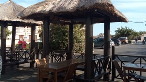 Une terrasse en bois avec des parasols en chaume portant l’inscription « PLAGE DE KUTA », des tables et des chaises en bois, et un bac à fleurs en pierre.