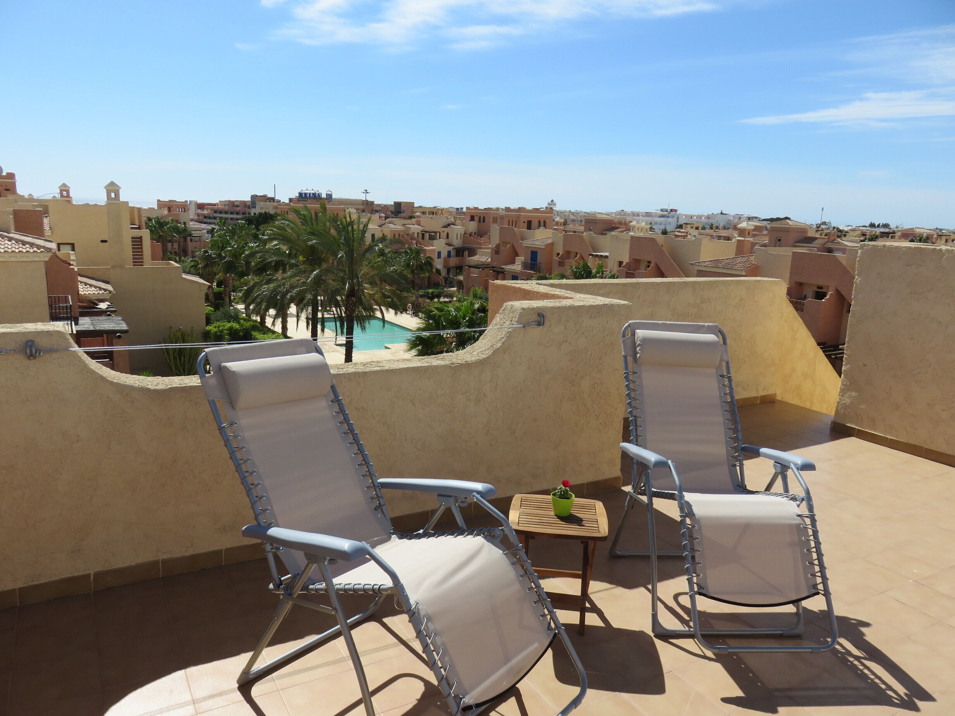 A rooftop terrace with two lounge chairs, a small table, and a view of a swimming pool and buildings in the distance.