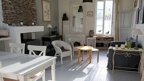 A spacious room with a high ceiling, white wooden flooring, and a stone wall. There are several white chairs, a table, and a sofa. A large window allows natural light to enter.