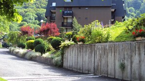 A multi-story residential building with a balcony on the second floor, surrounded by well-maintained gardens and a concrete retaining wall. - Studio 21m²- 2* - city center- wifi - balcony - private parking - linen included (Mont-Dore)