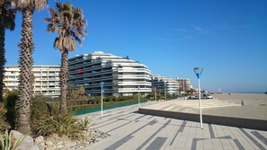 A beachfront promenade with modern buildings, palm trees, and a clear sky.