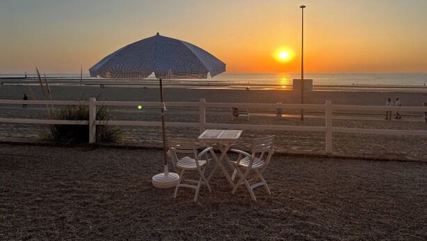 Et bord med stole og en parasol ved stranden, med udsigt over havet i solnedgang.