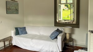 En hvid sofa med mønstrede puder, et glas sidebord, en lampe og et stort spejl der viser udsigt til en have.