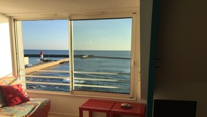 A room with a large window overlooking the sea, a red table, a TV, and a bed with colorful pillows. - Studio in house bordering the water, exceptional view over the harbour entrance (Guilvinec)