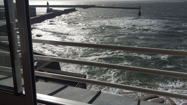 A view of the ocean from a ship's deck, with waves crashing against a pier and a lighthouse in the distance. - Studio in house bordering the water, exceptional view over the harbour entrance (Guilvinec)
