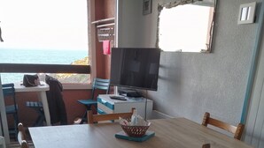A dining area with a wooden table and chairs, a television on a stand, and a view of the sea through a window.