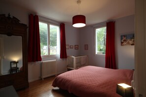 Ein Schlafzimmer mit einem großen Bett, einer hölzernen Kommode, einem Babybett und einem Fenster mit roten Vorhängen.