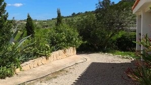 Un sentiero sterrato conduce a un edificio bianco con tetto in tegole, circondato da vegetazione e un muro in pietra.