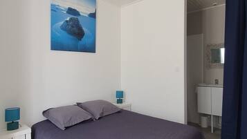 Un dormitorio con una cama, dos mesitas de noche con lámparas, un cuadro de un paisaje y un baño visible a través de una puerta abierta.